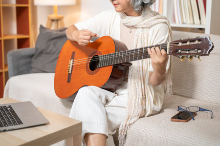 A close-up of a relaxed Asian senior woman practices an acoustic guitar in the living room while studying an online lesson through her laptop at home. leisure, lifestyles, retirementの写真素材