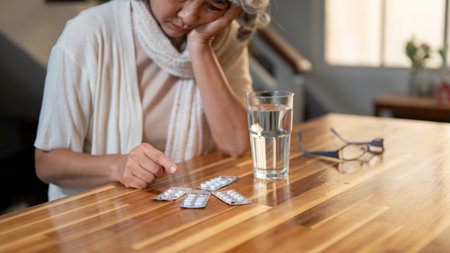 A close-up of a sad, bored elderly Asian woman looks at her medicines on the table with an unhappy expression, feeling sick and unwell, and bored of taking so many medicines.の写真素材