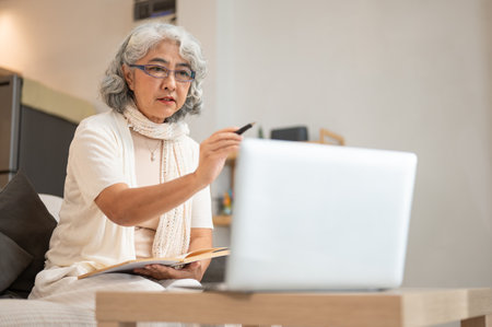 A positive Asian senior woman focuses on reading an article on the internet through her laptop while sitting on a sofa at home. people and technology conceptsの写真素材
