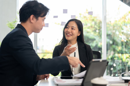 Two businesspeople are meeting in a meeting room, having a nice conversation. A beautiful, positive Asian businesswoman is working with her male colleague, discussing a project.の写真素材