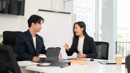 Two businesspeople are meeting in a meeting room, having a nice conversation. A beautiful, positive Asian businesswoman is working with her male colleague, discussing a project.の写真素材