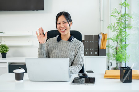 A beautiful, friendly Asian businesswoman sits at her desk in the office, waving and smiling at the camera. businesspeople conceptの写真素材