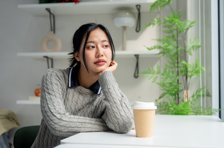 A beautiful Asian woman in a cosy sweater sits at a table in a minimalist coffee shop with a coffee cup on the table, looking out the window as she pondering something.の写真素材