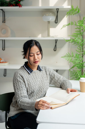 A beautiful, calm Asian woman in a cozy sweater relaxes while reading a book at a table by the window in a minimalist cafe. people and lifestyle conceptsの写真素材