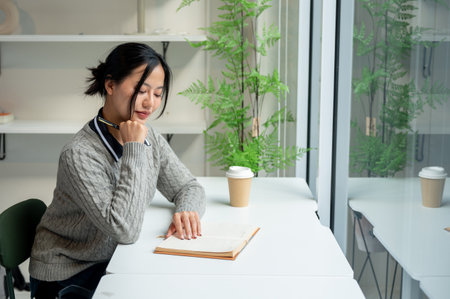 A beautiful, calm Asian woman in a cozy sweater relaxes while reading a book at a table by the window in a minimalist cafe. people and lifestyle conceptsの写真素材
