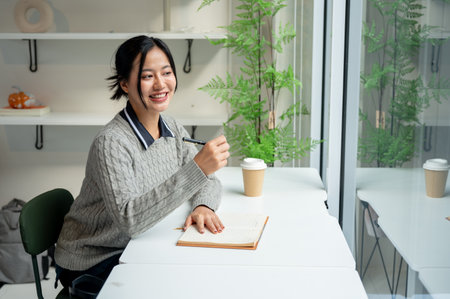 A positive, charming Asian woman in a cosy sweater sits at a table by the window with her diary placed on the table, smiling away.の写真素材