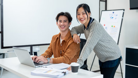 A portrait of two positive Asian businesspeople, a male and female, both are in the office and smiling at the camera. One sits at a desk in front of a laptop, while the other stands beside.の写真素材