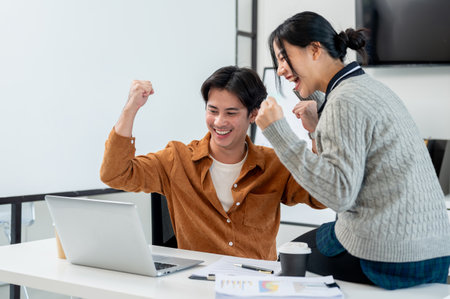 Two cheerful Asian businesspeople raise their triumph fists while looking at a laptop screen, celebrating their project success together in the office.の写真素材