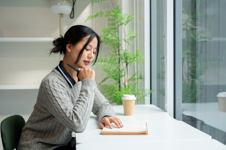 A beautiful, calm Asian woman in a cozy sweater relaxes while reading a book at a table by the window in a minimalist cafe. people and lifestyle conceptsの写真素材