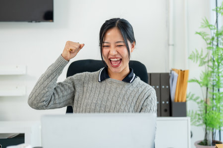 A cheerful, excited Asian businesswoman sits at her desk in the office, raising her fist in triumph and screaming with joy while looking at her laptop screen, celebrating her success or good news.の写真素材