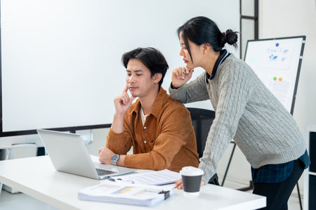 Two focused and serious Asian businesspeople look at a laptop screen, brainstorming solutions to fix the problems in their project, working together in the office.の写真素材