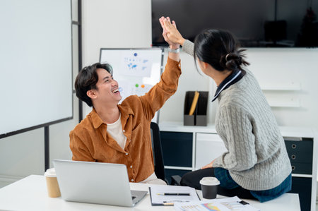 Two cheerful and happy Asian businesspeople are in the office, giving each other a high five to celebrate their project's success. teamwork, colleague, co workingの写真素材