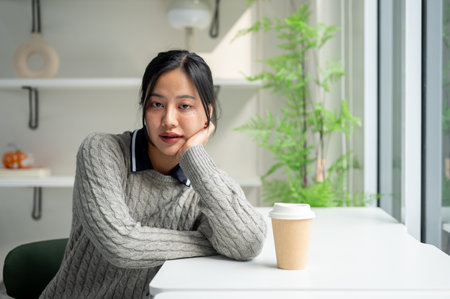 A charming Asian woman in a cosy sweater sits at a table in a minimalist coffee shop with a coffee cup on the table, looking at the camera.の写真素材