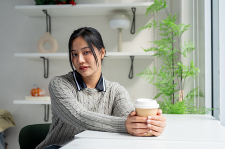 A beautiful Asian woman in a cosy sweater sits at a table in a minimalist coffee shop with a coffee cup on the table, looking at the camera.の写真素材
