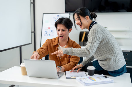 Two cheerful, positive Asian businesspeople brainstorm and discuss a project together in a modern office, looking at a laptop screen while conversing.の写真素材