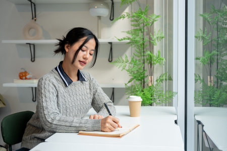 A beautiful, calm Asian woman in a cosy grey sweater writes in her diary while relaxing in a minimalist cafe. people and lifestyle conceptsの写真素材