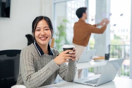 A charming Asian businesswoman in a cozy sweater sits at her desk in the office with a coffee cup in her hand, smiling at the camera. corporate job, marketing agency, employeeの写真素材