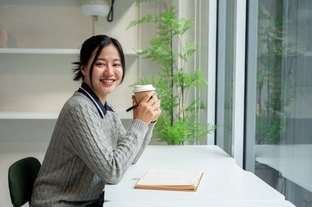 A positive, charming Asian woman in a cosy sweater sits at a table by the window with her diary placed on the table, smiling at the camera.の写真素材