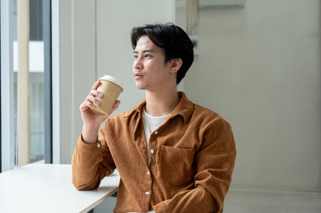 A thoughtful Asian man gazes out the window while sipping coffee in a minimalist cafe. people and lifestyles conceptsの写真素材