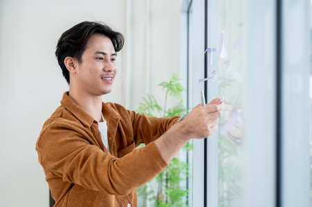 A positive, handsome Asian businessman attaches sticky notes on a glass window in the office, listing his ideas for a project while brainstorming with his colleagues.の写真素材