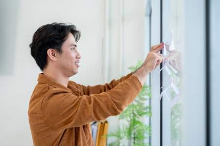 A positive, handsome Asian businessman attaches sticky notes on a glass window in the office, listing his ideas for a project while brainstorming with his colleagues.の写真素材