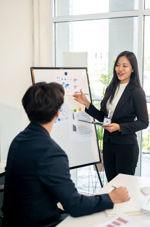 A confident, positive Asian businesswoman is presenting her project to her team in a meeting room, explaining charts on a board. business people and meeting conceptsの写真素材