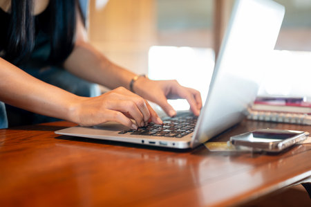 A close-up of a woman working remotely on her laptop in a coffee shop, fingers typing on the laptop keyboard. people and technology conceptsの写真素材