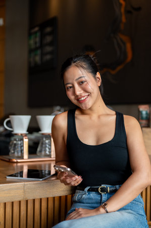 An attractive Asian woman sits at a counter bar in a coffee shop, holding her smartphone and smiling at the camera. people, lifestyles, technologyの写真素材