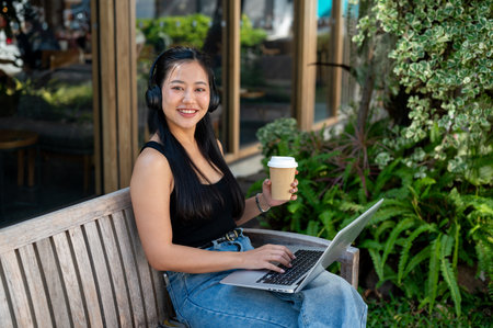 A confident Asian woman is working remotely outdoors, sitting on a bench in a garden, working on her laptop and smiling at the camera.の写真素材