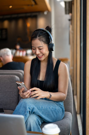 A happy, smiling Asian woman enjoys listening to music on her headphones while chatting with her friend on her smartphone in a cafe. people, wireless technology, lifestylesの写真素材
