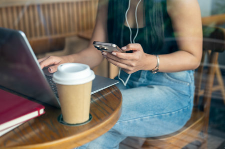 A cropped image of a beautiful Asian woman wearing earphones, using her smartphone while working remotely from a coffee shop in the city. people and technologyの写真素材