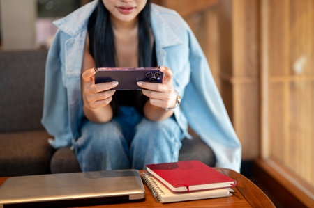 A close-up of a woman in a denim jacket sits in a coffee shop, using her smartphone, watching videos or playing a mobile game. people and wireless technologyの写真素材