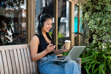An Asian woman wearing headphones is talking on a video call or online meeting through her laptop while sitting on an outdoor bench, working remotely.の写真素材