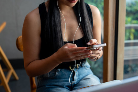 A cropped image of a beautiful Asian woman wearing earphones, using her smartphone while working remotely from a coffee shop in the city. people and technologyの写真素材