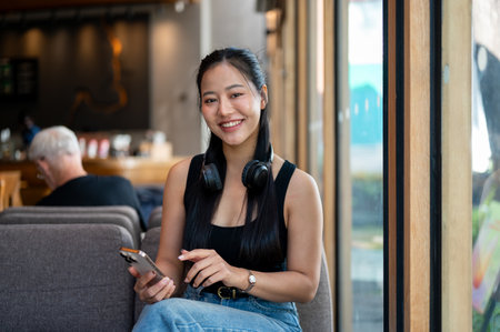 An attractive Asian woman in casual clothes sits on a sofa in a coffee shop, holding a smartphone in her hand and smiling at the camera. people and lifestyles conceptsの写真素材