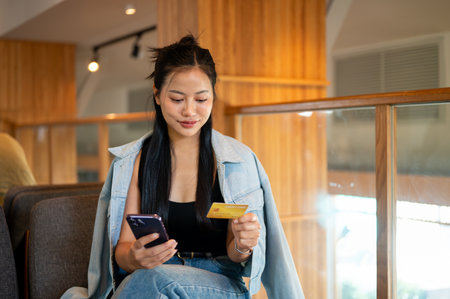 A beautiful Asian woman sits in a contemporary coffee shop, holding her smartphone and a credit card, shopping online. online payment, mobile banking, e-commerce, lifestylesの写真素材