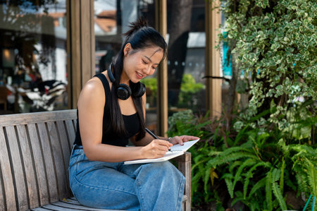 A beautiful, contemplating Asian woman writing her diary while relaxing on an outdoor bench in a garden. leisure, hobby, lifestyleの写真素材