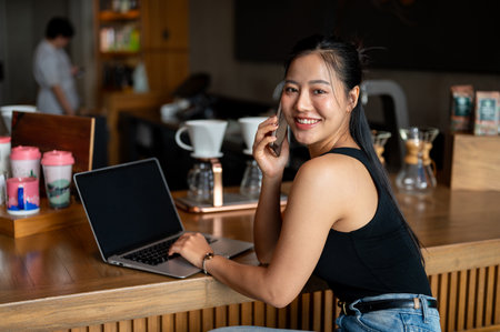 An attractive Asian woman sits at a counter bar in a coffee shop with her laptop, working remotely, talking on the phone and smiling at the camera.の写真素材