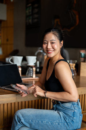 An attractive Asian woman sits at a counter bar in a coffee shop with her laptop, working remotely, holding her smartphone and smiling at the camera.の写真素材