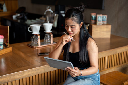A beautiful Asian woman sits at a counter bar in a coffee shop with a digital tablet in her hand, contemplating while looking at the tablet screen, working remotely.の写真素材