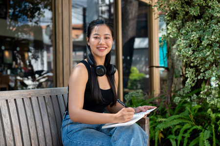 A portrait of a beautiful Asian woman in casual clothes sitting on an outdoor bench in a garden with her diary, smiling at the camera. leisure, hobby, lifestyleの写真素材