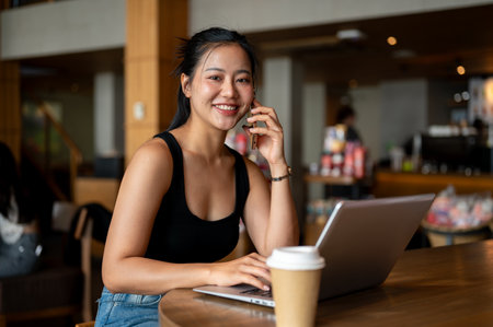 A confident, attractive Asian woman is talking on the phone while working remotely from a coffee shop, smiling at the camera. businesswoman, freelancer, digital nomadの写真素材