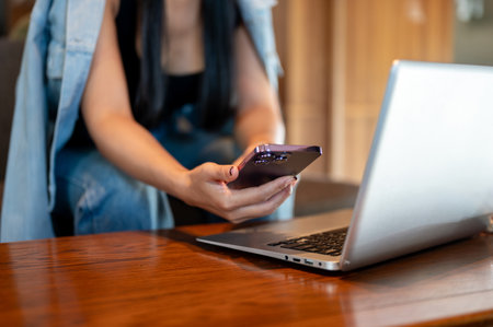 A close-up of an Asian woman in casual clothes using her smartphone while working remotely on her...