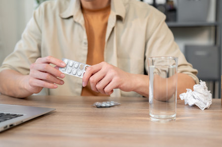 A close-up of an unwell businessman sitting at his desk in the office, holding a packet of pills in his hand and struggling with sickness while trying to work. health care, medical, sicknessの写真素材