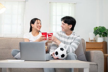 A lovely, carefree young Asian couple, boyfriend and girlfriend, enjoy drinks while watching a football game together at home. bonding, sport game, leisureの写真素材