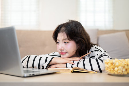 A young Asian female university student is resting her chin on her hands in front of her laptop at a...