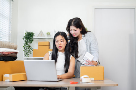 A beautiful Asian woman is showing e-commerce shipping orders on the website on a laptop to her colleague while preparing items for shipping, working together in the office. e-commerce, online shopの写真素材