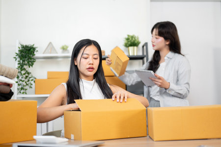 A beautiful, positive Asian female stock inventory manager is packing client orders and checking them on her laptop, while her colleague is reviewing stock items on shelves in the background.の写真素材