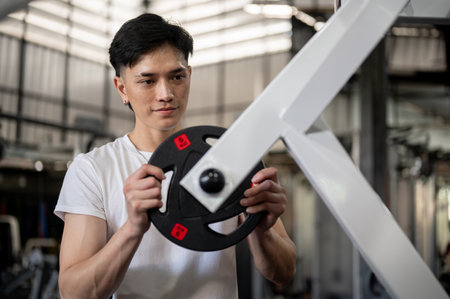 An Asian man placing a weight plate on a machine, preparing it to train his muscles while working out in the gym. healthy lifestyle, body building, muscular training, weight liftingの写真素材