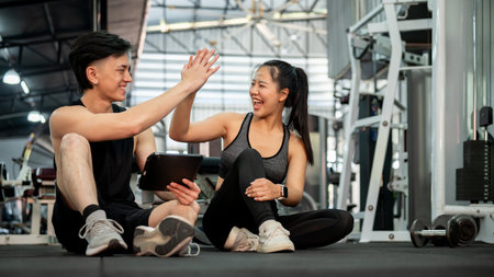 A cheerful Asian male fitness trainer and his female client are sitting on the floor in the gym, giving each other a high five after finishing their workout session. fitness life, healthy lifestyleの写真素材